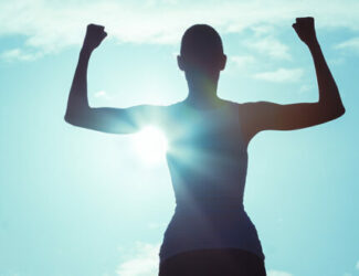 Silhouette of a person standing outdoors with arms raised in a triumphant pose against a bright, cloudy sky, with sunlight shining through, symbolizing strength and positivity